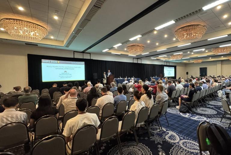 The IMPS conference, featuring people watching speakers on a stage at the back of the room.