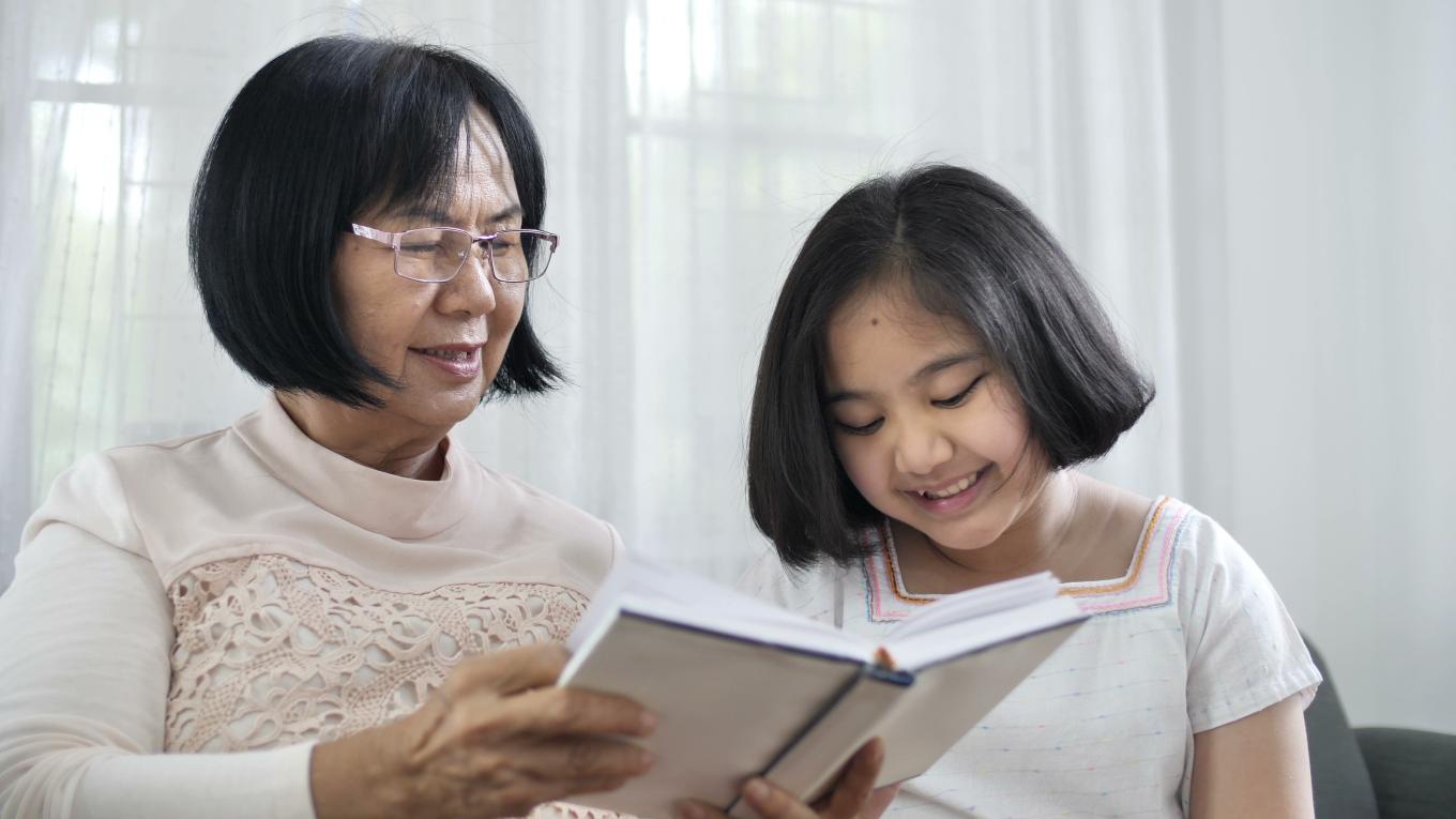 Adult sitting on a couch and reading a book to a smiling child.