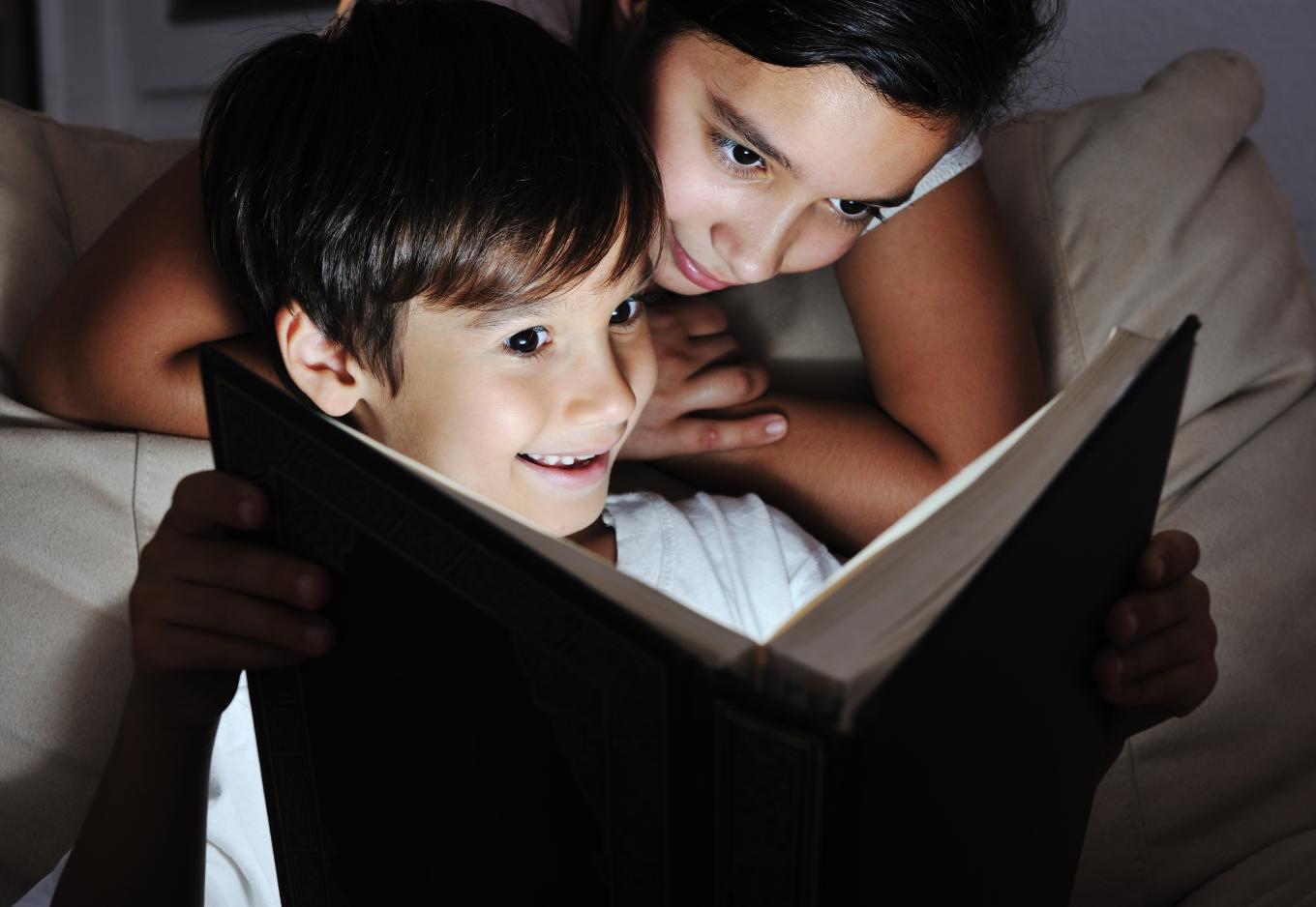 Two children reading in the dark. One child is on a couch or bed looking at the book over the other child's shoulder.