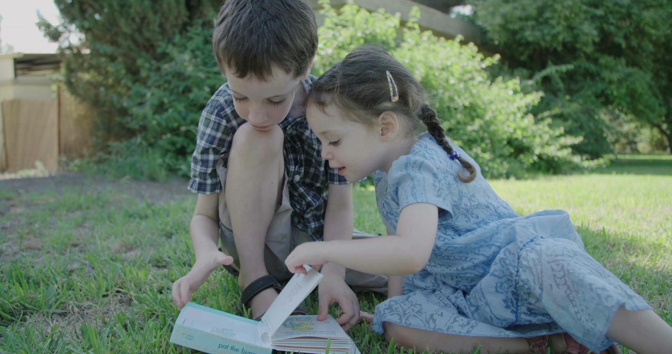 Two children sitting on the grass and reading a book outside. One child is turning a page.