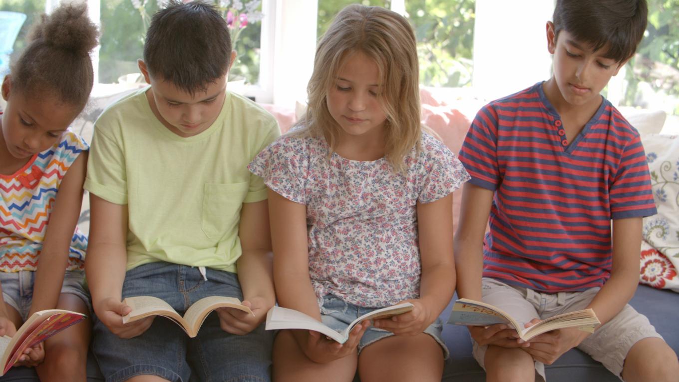A group of four children sitting side-by-side in front of windows and reading.