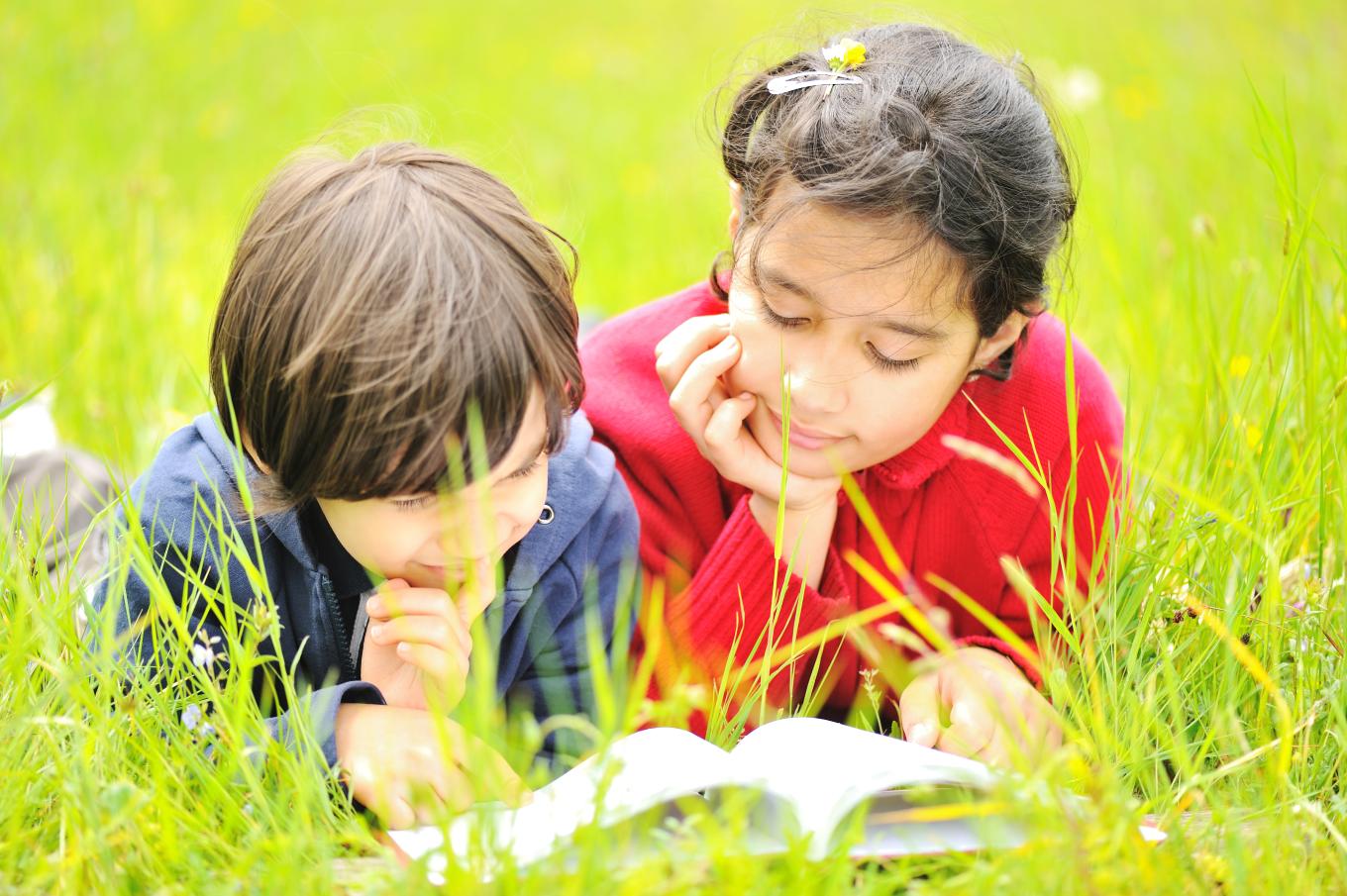 Two children laying in the grass outdoors and reading a book together.