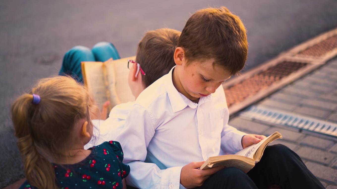 Three children sitting with their backs to each other reading books on the sidewalk outdoors next to the pavement.